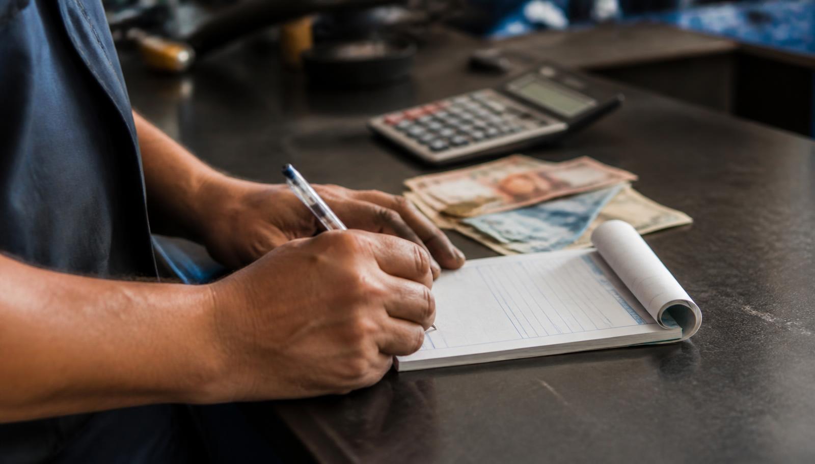 Técnico preenchendo um bloco de recibo de pagamento na bancada, com calculadora e dinheiro ao lado