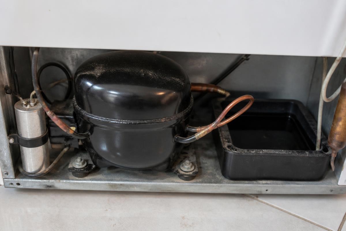 Close da base traseira de uma geladeira com o compressor preto parafusado e a bandeja plástica de água ao lado