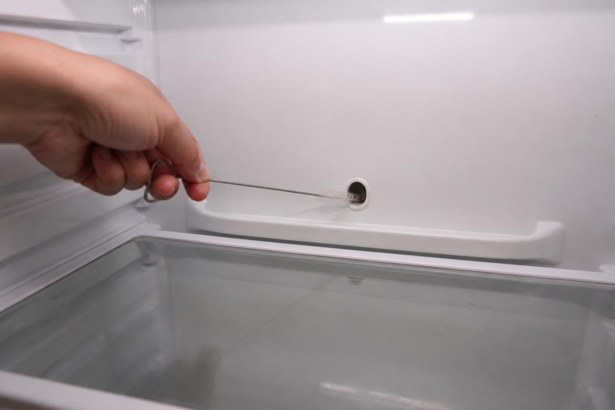 Mão passando uma escova fina e comprida dentro do furo do dreno de degelo no fundo interno de uma geladeira