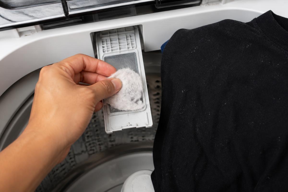 Filtro de fiapos da lavadora sendo limpo ao lado de roupa preta com fibras brancas aparentes