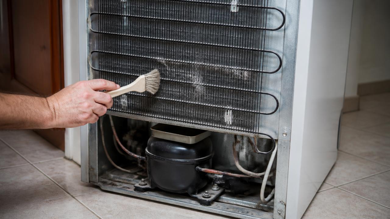 Parte traseira de geladeira com condensador coberto de poeira sendo limpo com escova, compressor preto visível na base