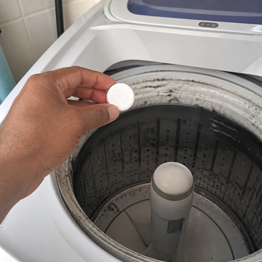 Mão segurando pastilha efervescente branca de limpeza sobre o cesto vazio de máquina de lavar de abertura superior